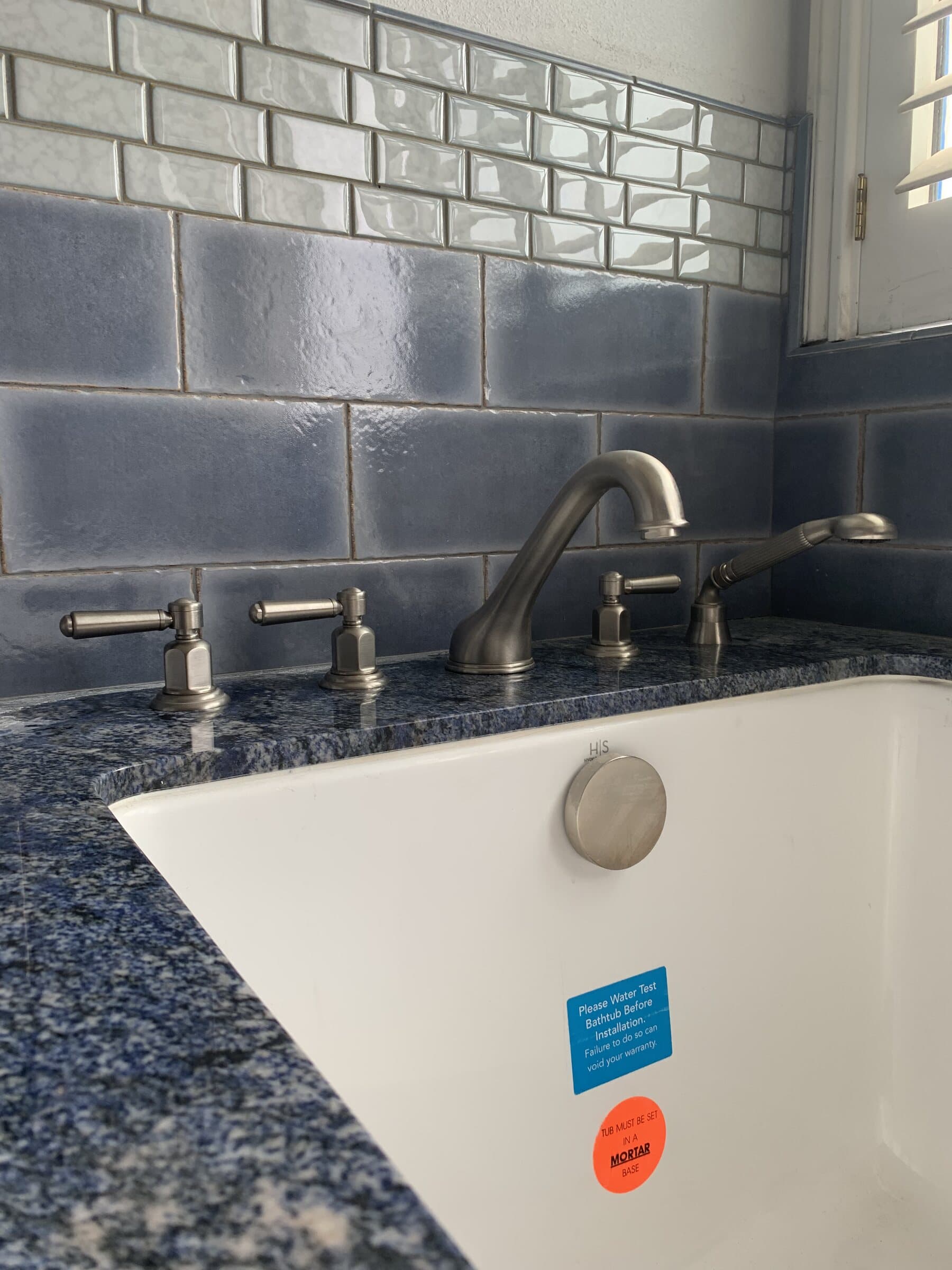 Deck-mounted tub filler with cross handles and a hand shower on a dark granite tub deck, with a grey subway tile surround
