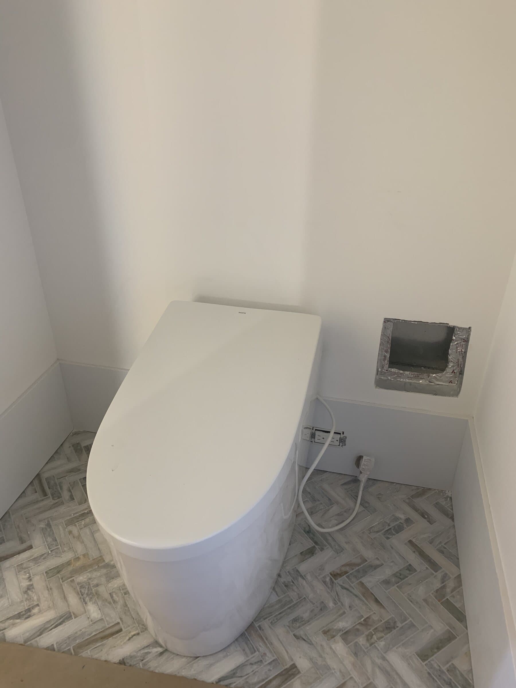 Skirted one-piece electronic bidet toilet on a herringbone marble tile floor, with a wall-mounted control panel and a dedicated receptacle beside it
