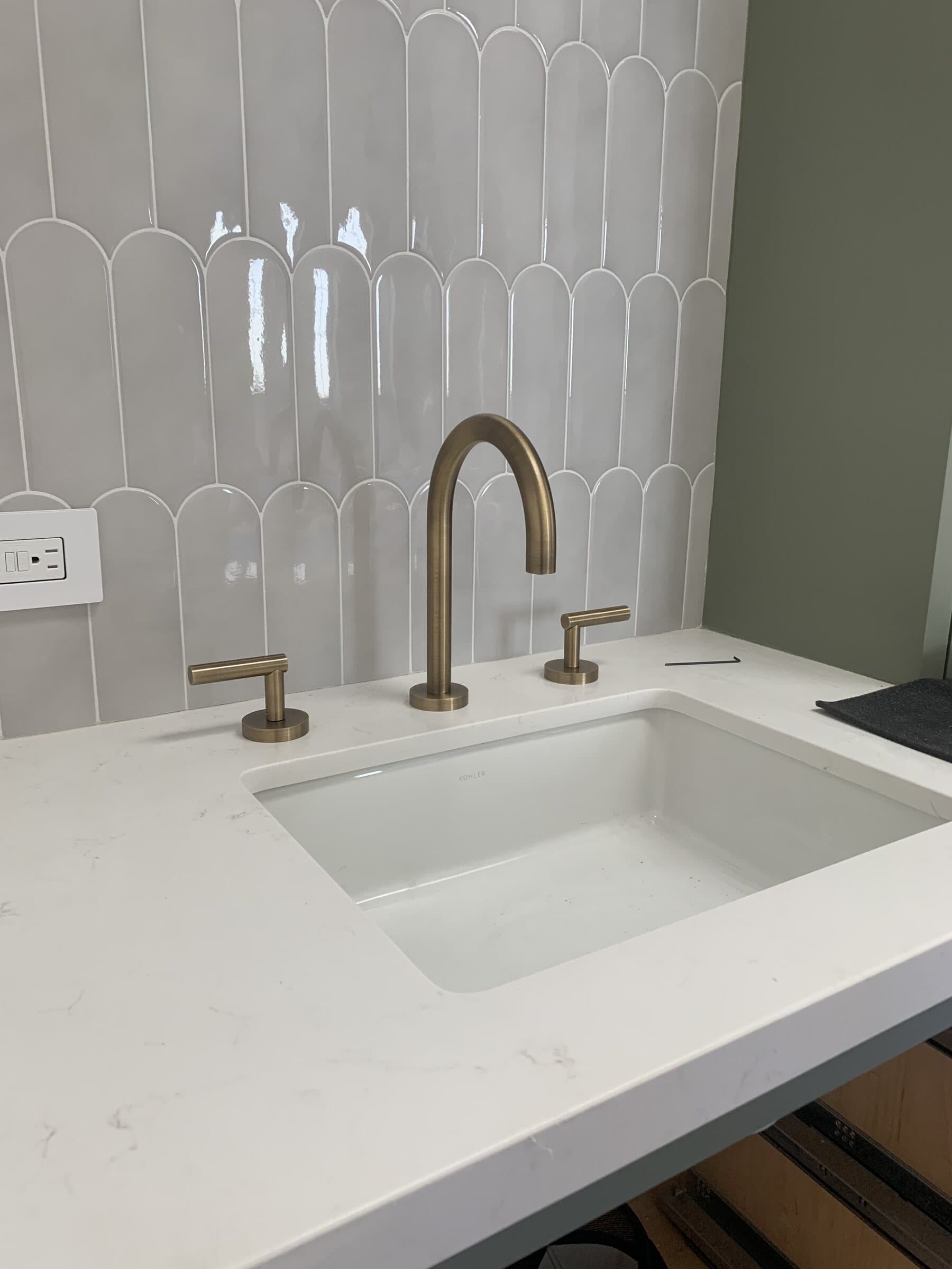 Undermount sink in a white quartz vanity top with a brushed brass gooseneck faucet against a white fluted tile backsplash
