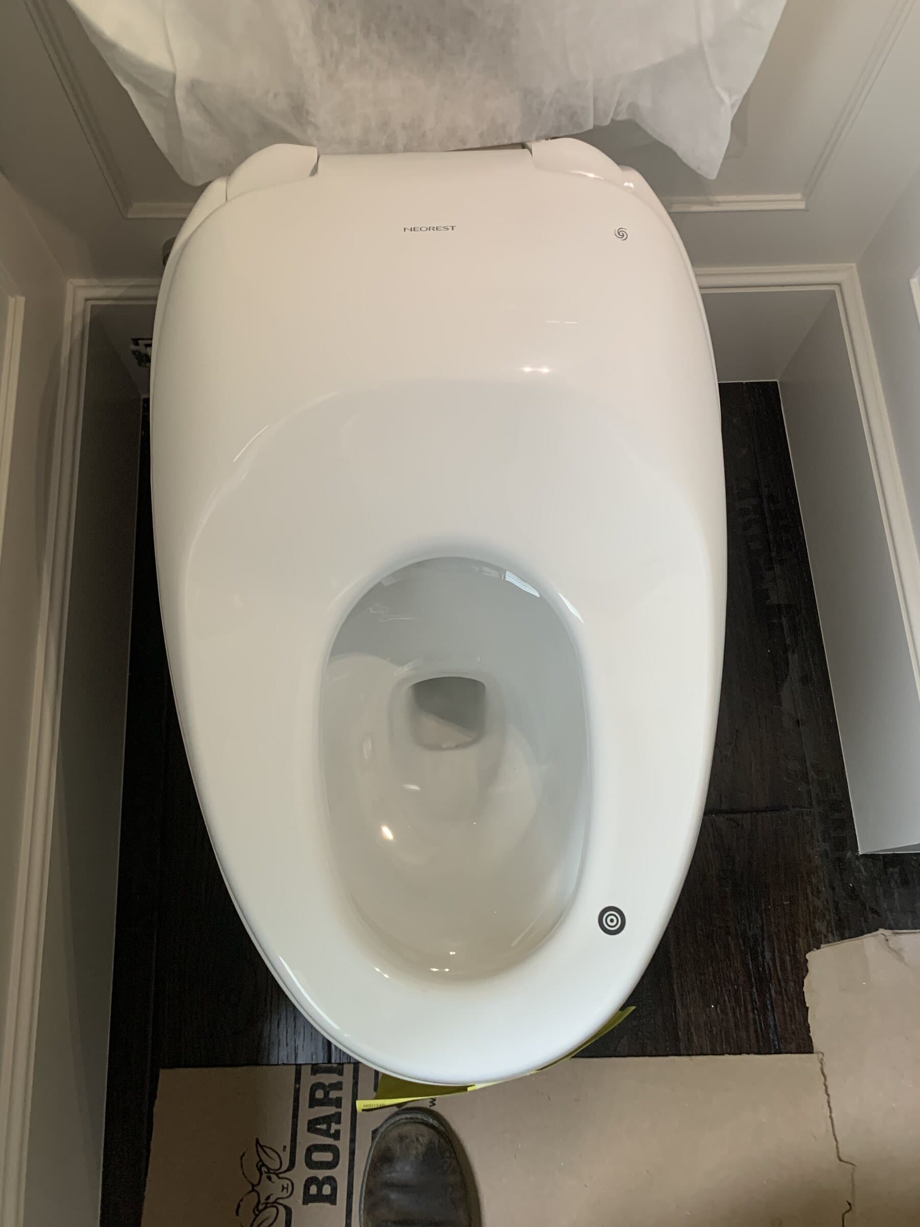 Skirted one-piece toilet being set in a bathroom under construction, with protective sheeting overhead and the finished floor covered