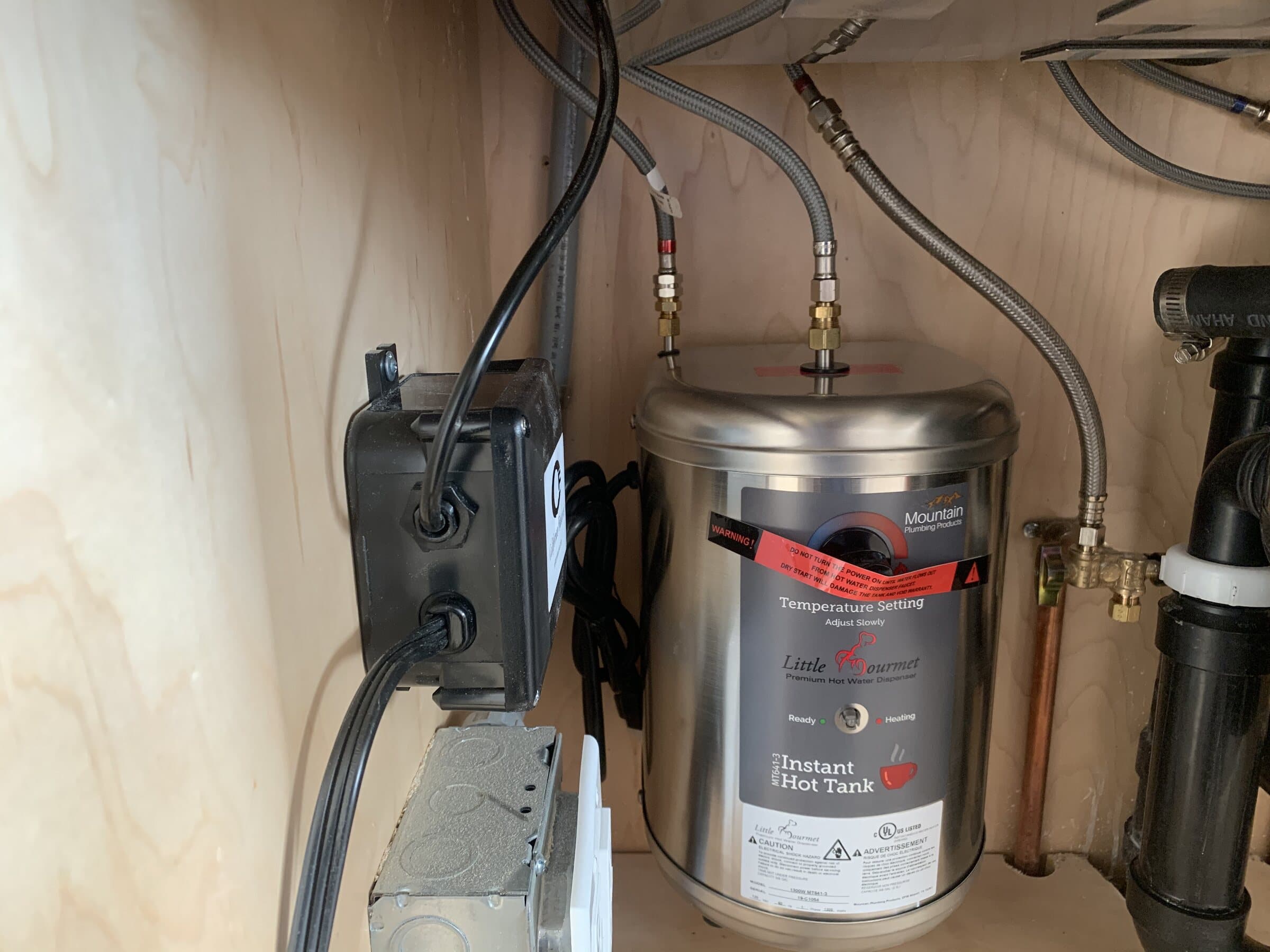 Stainless steel instant hot water tank mounted under a sink with braided supply lines, a transformer and copper connections