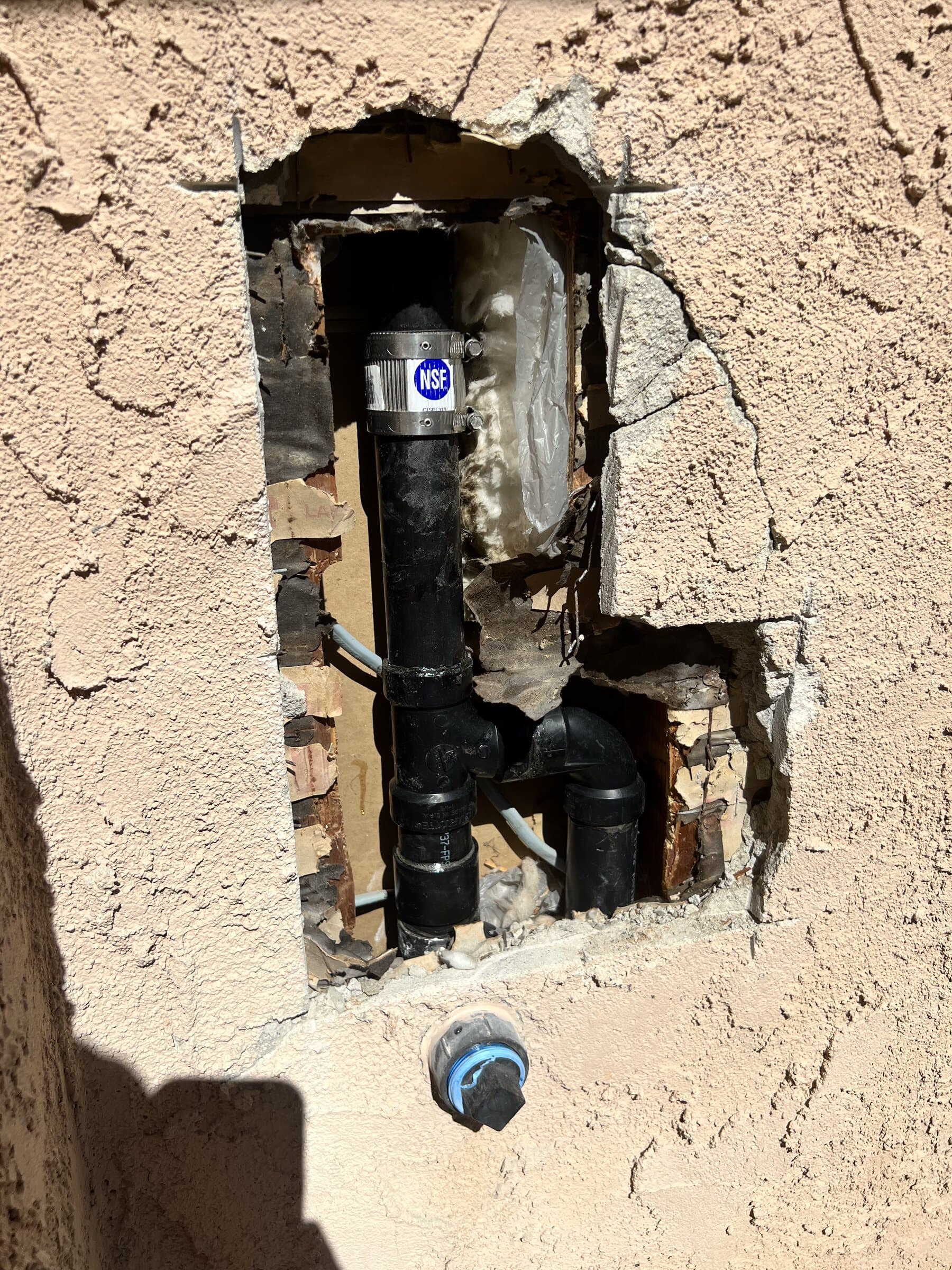 Stucco exterior wall cut open to expose black ABS drain piping and a new cleanout fitting during a sewer repair