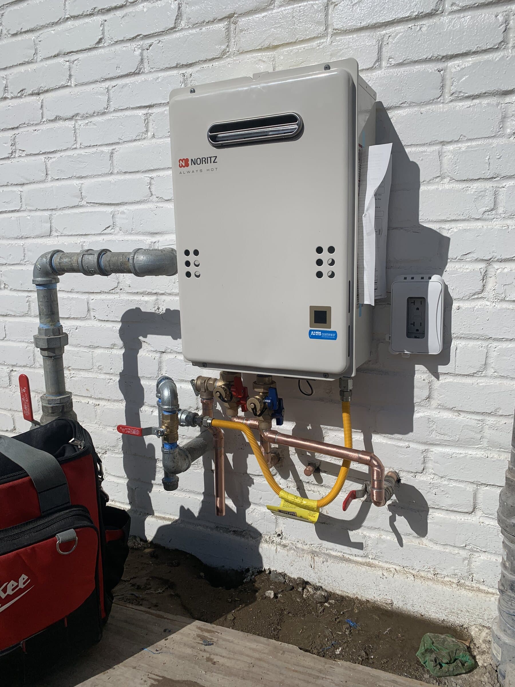 Noritz tankless water heater mounted on a whitewashed brick exterior wall with galvanized gas piping, a yellow flexible gas connector and copper water lines