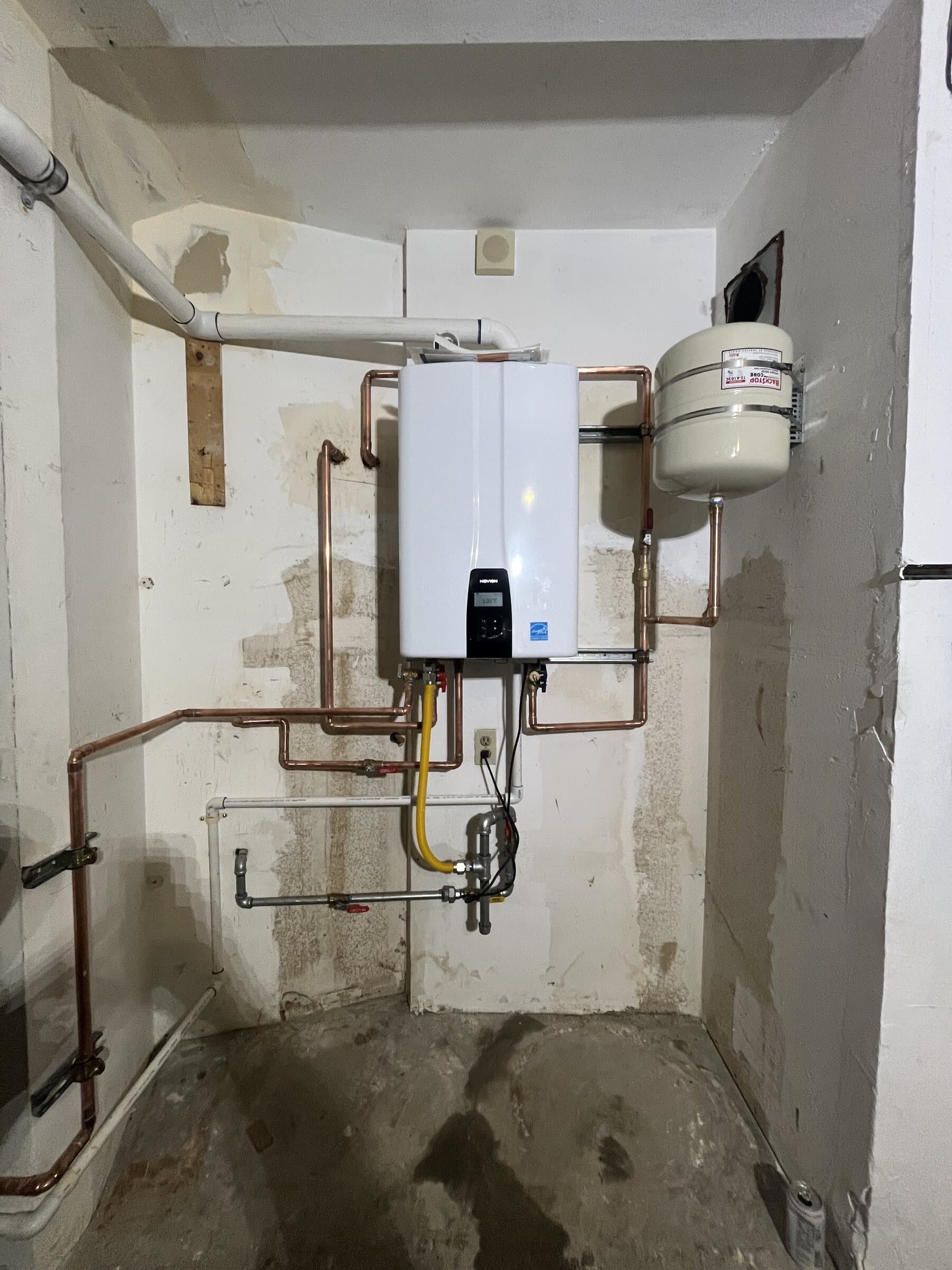 Wall-mounted tankless water heater in an unfinished utility space with copper supply lines, a gas shutoff and a condensate line