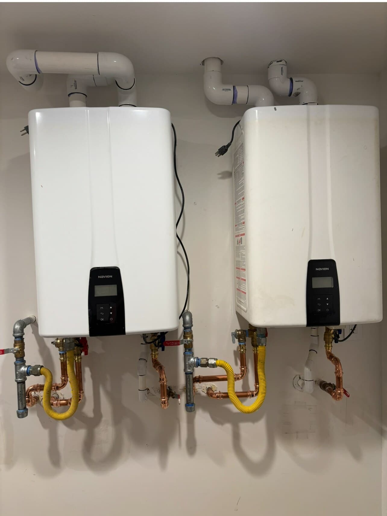 Two wall-mounted Navien tankless water heaters installed side by side with PVC venting, yellow CSST gas connectors, copper water lines and isolation valves